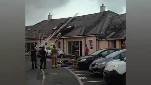 Firefighters are using a long ladder to access the roof of a block of flats and damage can bee seen to roofing tiles.