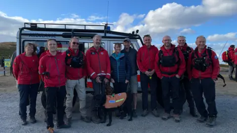 Mountain rescue volunteers and search dogs standing in front of a rescue vehicle for a group photo.