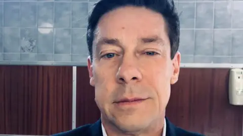 BBC A mirror selfie of a man in a bathroom with blue tiles. He is in his mid-50s and has a half-smile expresssion. Although the photo is cropped you can tell he has a suit, shirt and tie on and is freshly shaven.