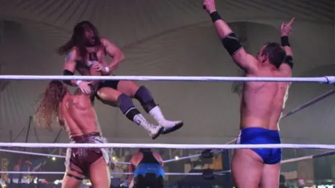 Four wrestlers inside a ring. One with his back to us is lifting another in the air around the throat and is about to slam him down. Another man is on the right with both hands raised in the air. A fourth man is slumped over the ropes on the far side. 