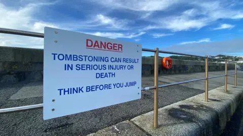 Guernsey Coastguard A sign attached to a railing near a sea wall that reads: Danger, tombstoning can result in serious injury or death, think before you jump. In the background there is a life ring attached to the sea wall.