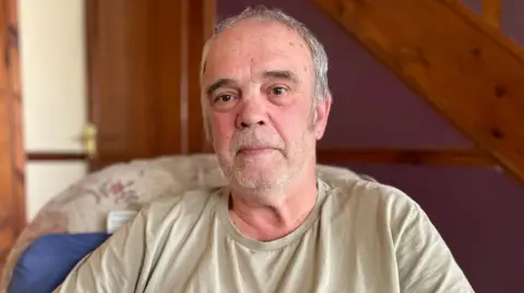 A man with thinning white hair wearing a khaki T-shirt looks sadly at the camera.