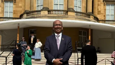 A man smiling in a dark suit, whit shirt and purple tie standing proudly on a lawn in front of a grand building. Several other smartly-dressed people are nearby under a white canopy at the building's entrance. 
