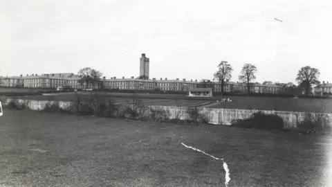 A black and white photo showing a large asylum building with a central tower and surrounded by trees and grass