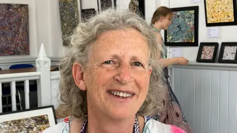 Luke Deal/BBC Vivica Bamford stands in the library's gallery with The Hat's artwork on display behind her. She has short, curly grey hair and wears a white top with some floral details on it.