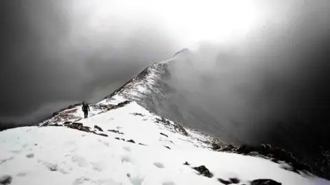 Lee Gilchrist A man is tiny walking along the ridge of a snowy mountain. The picture is black and white.