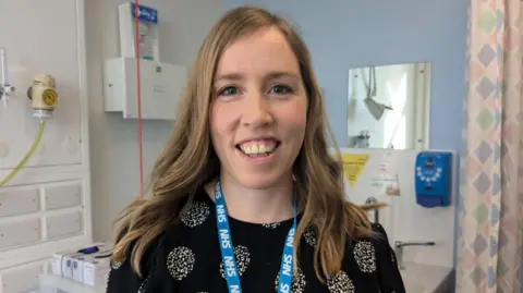 Kit Taylor/BBC A woman with shoulder length wavy blonde hair smiling at the camera. She is wearing an NHS lanyard and in the background is a hospital check-up room. 