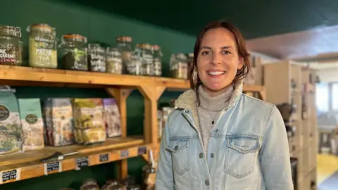 Jess stands in front of shelves filled with various jars and packages in a store. Shel is wearing a light denim jacket over a turtleneck sweater. The shelves behind them contain glass jars labeled with different types of tea and other items, as well as some packaged goods. The background shows more shelving units and the interior of the store.