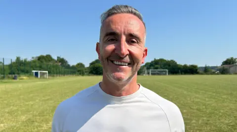 BBC/Gavin Kermack A man with short greying hair and grey stubble wearing a white t-shirt and standing on a football pitch, with a goal in the distance. He is smiling at the camera.