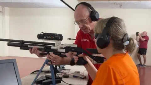 Una sesión de tiro al blanco en interiores, donde un instructor ayuda a un participante a utilizar un rifle de aire comprimido de precisión.