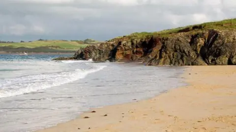 Part of Tregirls Beach near Padstow in Cornwall. The waves are crashing into the sand. A craggy area of the coast is in the background.