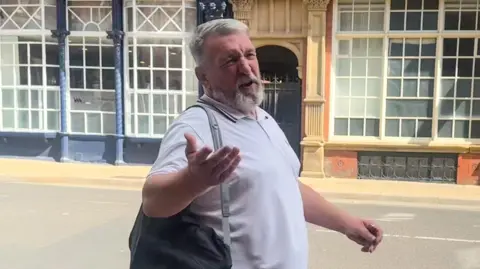 A man with short grey hair and a beard stands on a street, wearing a light polo shirt and a dark shoulder bag, gesturing with one arm in front of a building with large multi‑paned windows and ornate blue‑and‑beige architectural details.