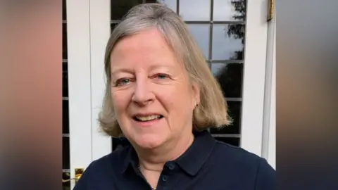 RNLI A blonde woman. She is wearing a black polo shirt and is standing in front of a white window. 