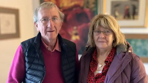 peter, left, has short grey hair. He is wearing thin framed glasses and a black body warmer. He is wearing a raspberry coloured jumper and a pink shirt underneath. Susan, right, has shoulder length blonde hair and is wearing brown glasses. She has a purple coat on and a red animal print shirt underneath.