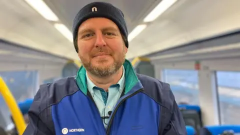Paul is wearing a Northern uniform, standing in a railway carriage, smiling at the camera.
