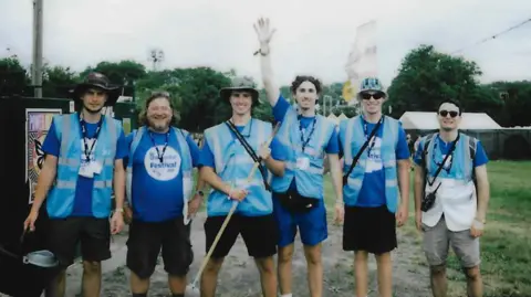 WaterAid A film photo showing a group of six WaterAid volunteers wearing hi-vis branded blue vests and lanyards. They are carrying equipment like a mop and a bucket to clean toilets. They are smiling and posing for a photo.