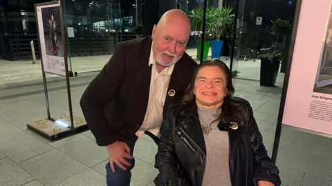 Phil Chapman / BBC A bald man crouches next to a woman in a wheelchair, both are looking at the camera, pictured in an auditorium 