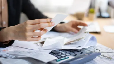 A white hand holds a receipt over a table containing other receipts, a calculator and pair of glasses