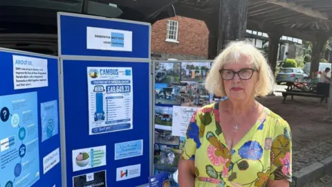 A woman with medium length blond hair and glasses wears a patterned yellow dress and glasses. She is standing next to a promotional board infront of an ancient market square.