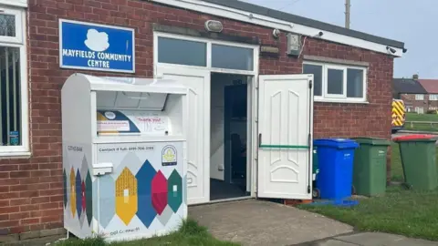 Tyesha Muncaster A single storey brick building with white double doors open and a sign on the wall saying Mayfields Community Centre. 