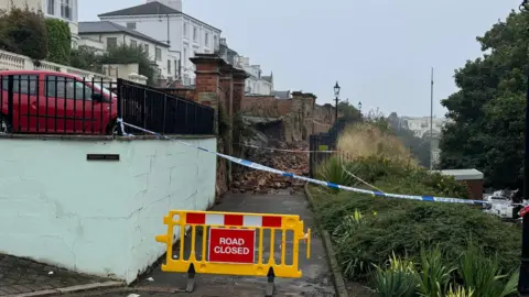 A 'road closed' sign blocking a pathway where a wall has collapsed. 