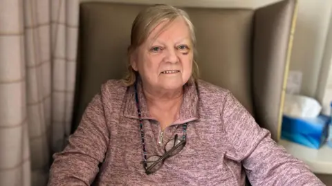 A woman wearing a lilac coloured top, with a zip and collar, sitting in a chair looking into the camera. She has glasses on lace around her neck and has short blonde hair