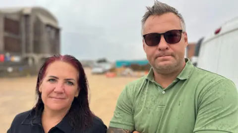 George Carden/BBC Liz Donnelly and Lee Smith stand on a beach near Shoreham seafront. Donnelly is wearing a dark top, while Smith is wearing sunglasses and a green polo shirt. Buildings and beach structures can be seen in the background.