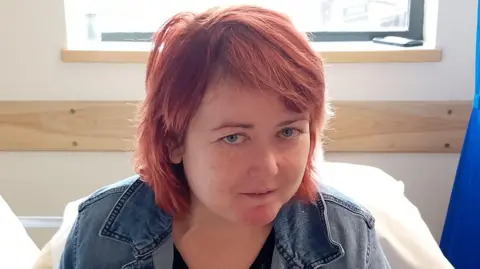 short red hair and is wearing a black top under a blue denim jacket. She is sitting on a bed in a hospital or clinic's room, two large white pillows are sitting behind her. Further back is a window.