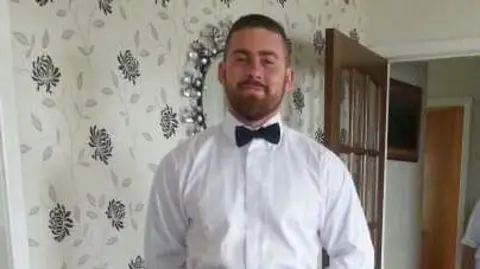 Kevin McCauley Frazer Irvine stood in a room which has flowery wallpaper. He has a brown beard and short hair. He is wearing a white shirt and black bowtie. A wooden door behind him is open.