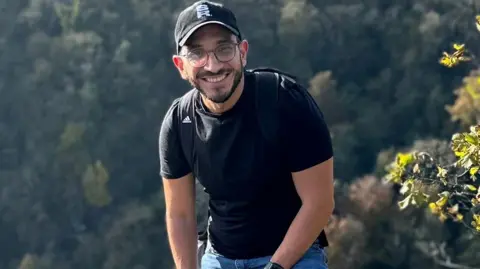 Roger Munoz Roger Munoz has a black T-shirt on and blue jeans and is wearing a black cap with S R in white block letters. He has glasses and a beard.