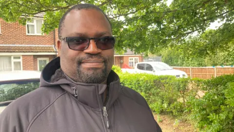 Mousumi Bakshi/BBC A man wearing a black coat and sunglasses is stood on a driveway with a house and greenery behind him