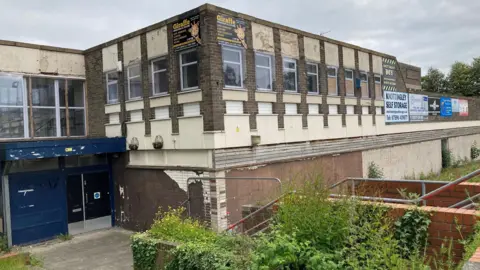 LDRS A visibly decrepit brutalist building with sharred paintwork. The grass around it looks overgrown.