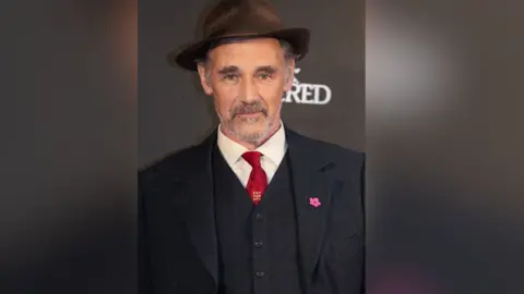 Sir Mark Rylance smiles into the camera wearing a navy pinstripe suit and waistcoat with a red tie and a brown fedora hat.