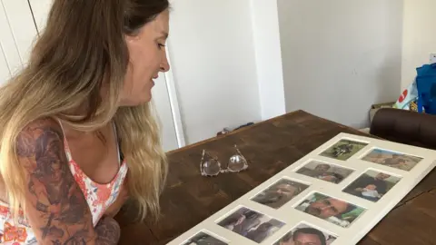 Caroline looks at a collage of photos featuring her husband Simon. The collage is resting on a table and Caroline is leaning on her arms looking down at it.