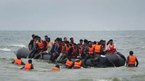 An overcrowded inflatable boat carrying dozens of people wearing orange life jackets sits in shallow coastal water. Several people are standing or wading alongside the vessel while others remain seated onboard. The sea is relatively calm, with a grey horizon and overcast sky in the background.
