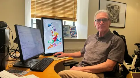 Professor Peter Hammon sits ebhind his desk which has two screens with maps and graphs of data.