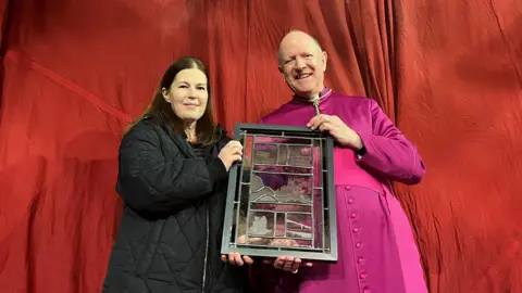 Jon Wright Danielle Hopkinson - a woman wearing a black jacket - presenting the Right Reverend Martin Seeley - a man wearing pink regalia - with a stained glass artwork. They are both standing in front of a red material backdrop 