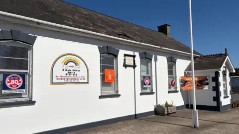 Deramore Orange Hall, a single-storey Victorian building painted white with a grey slate roof, to the right of the photo is a covered porch of the same construction and a flagpole stands in the centre of the image. Rainbow Corner's logo is on a sign in the centre of the wall.