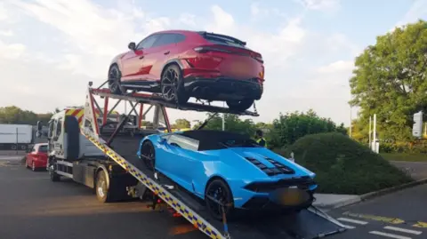 Two cars being towed away. One car is a blue sports car and the other car is red SUV. There are other cars on the road and a tree and shrubs to the right of the picture.
