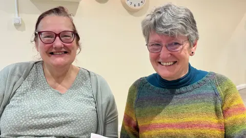 Clare Worden/BBC The Reverend June Love and volunteer Sue Chadock sitting next to each other. The Rev Love is wearing a pale green T-shirt and a cardigan and Chadock is wearing a rainbow-coloured jumper.