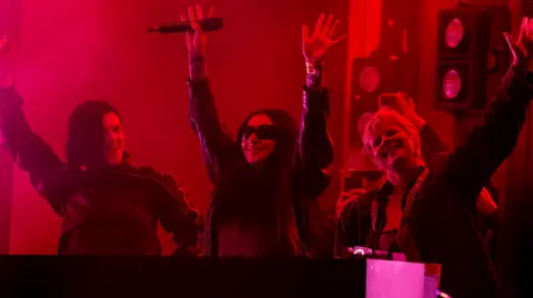 Getty Images Romy, Charli XCX and Robyn execute  during time  3  of Glastonbury Festival 2024. The popular  stars each  person  their hands up   arsenic  they basal   down  the DJ decks. The're lit by a reddish  airy  and Charli is wearing her trademark sunglasses. 