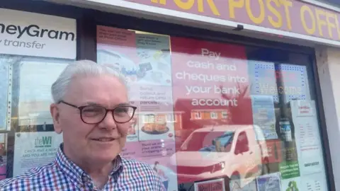 Annabel Amos/BBC Chris Attridge with short white hair and glasses looking at the camera. His post office is behind him, with several posters in the window and a poster saying "pay cash and cheques into your bank account". The reflection of a white van can be seen in the window.