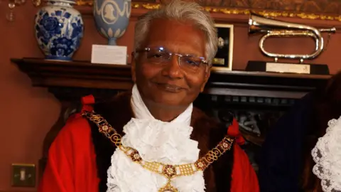 Wearing his mayoral robes and gold chain, Bharat Pankhania smiles at the camera. Behind him two vases and a musical instrument can be seen on a mantelpiece.