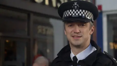 Jamie Niblock/BBC Insp Ben Jarvis, a Norfolk Police officer. He is standing outside in black and white uniform and is smiling.