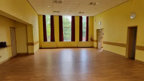 Wellingborough Town Council The inside of a building, showing a wooden floor, yellow walls, with three doors, a lock on the right hand side and three windows with the burgundy curtains. The room is empty. 