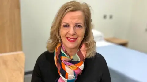 Caroline Donovan smiling while looking directly at the camera. She has long blonde hair and is wearing a black top and multicoloured scarf.