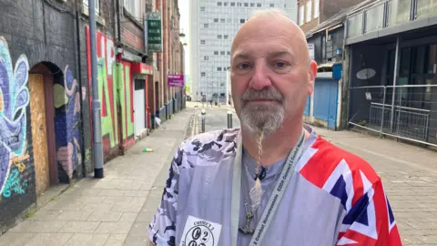 A man wearing a jacket that has a Union Jack on the left shoulder stands on a city centre street. He has a beard which has been plaited. There is graffiti on the walls of the buildings behind him and a large tower block in the distance.