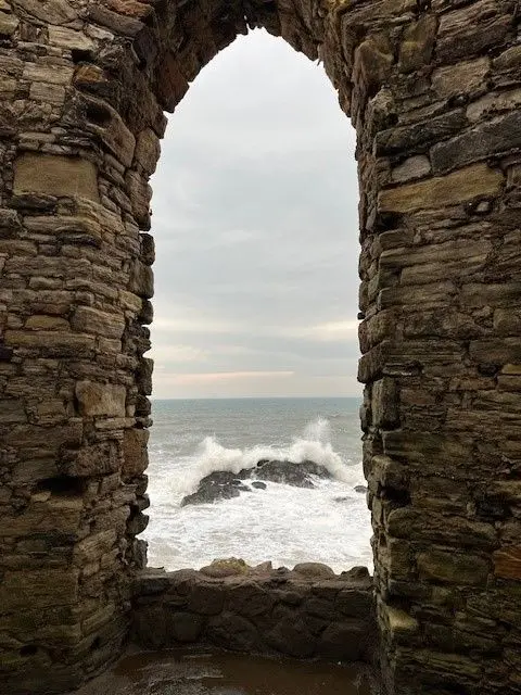 Gillian Watt waves crashing against a rock visible through a gap in a stone wall