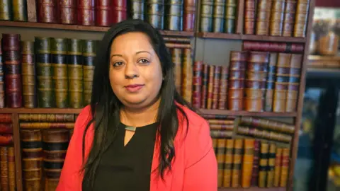 Michael Keohan/BBC A woman with black hair, wearing a tomato-red suit and black shirt, is standing in front of a book case. 