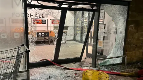 Large glass doors that have been smashed in, with a yellow defibrillator on the floor along side loads of glass.